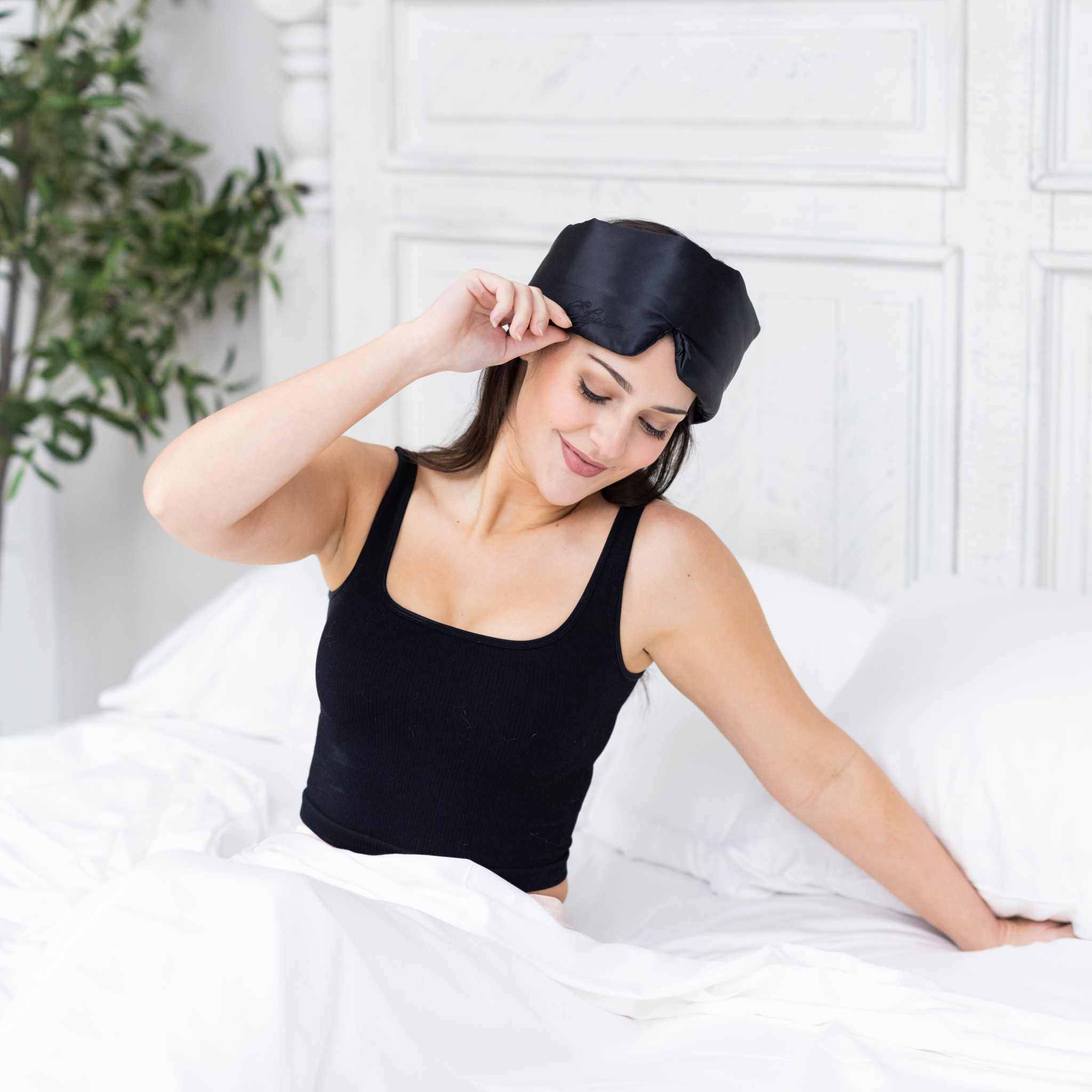 Young woman sitting in bed with Elysium Silk Eye Mask for sleeping in Black