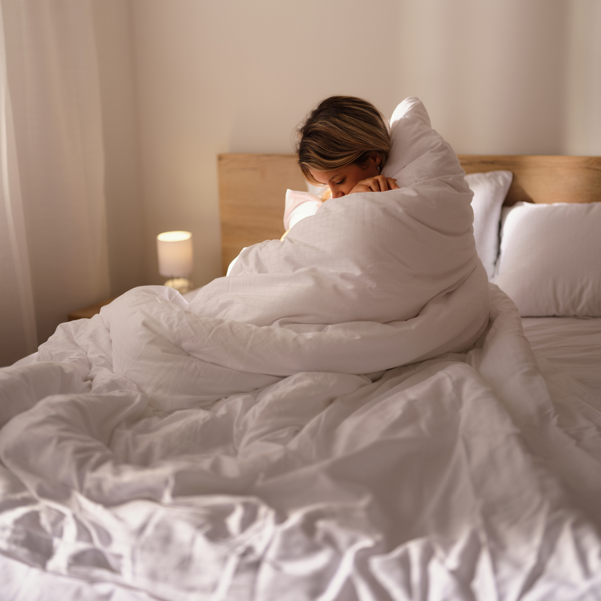 Young woman wrapped in a white comforter sitting on a bed in a softly lit bedroom