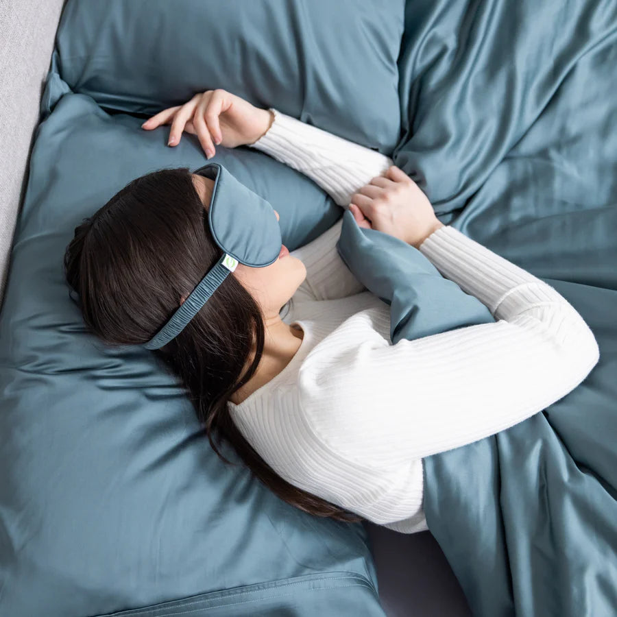 Woman sleeping on a bed with blue sheets, wearing a matching blue sleep mask and a white sweater